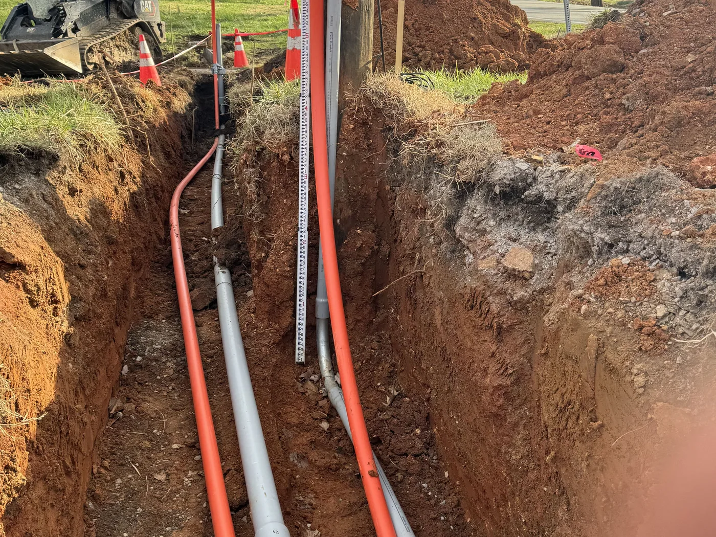 Orange and gray conduit runs laid in a deep utility trench, skid steer parked above